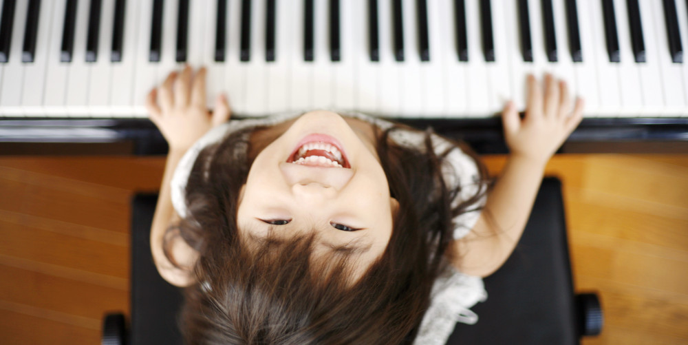 Featured image for "Piano Lessons Nashville TN" page on MusicLessonsNashville.com depicting girl approximately 5 years old playing the piano and looking up at the camera with a wide-mouthed smile.